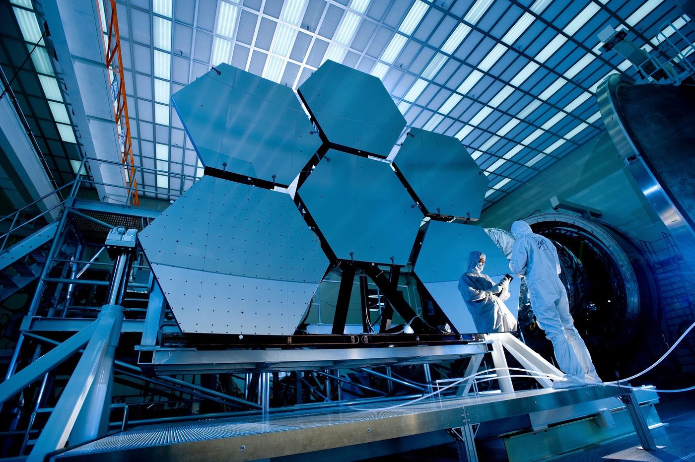 Cleanroom-suited engineers beside a hexagonal segmented telescope mirror under blue skylight