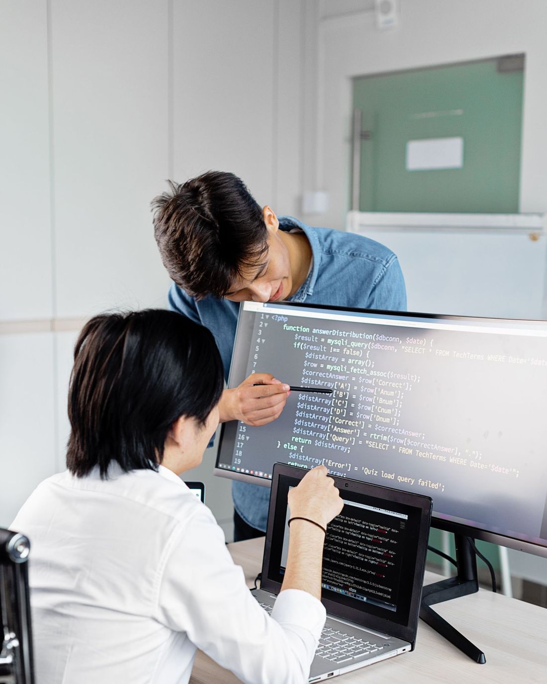 Two engineers reviewing code together — one pointing at a large display, the other at a laptop