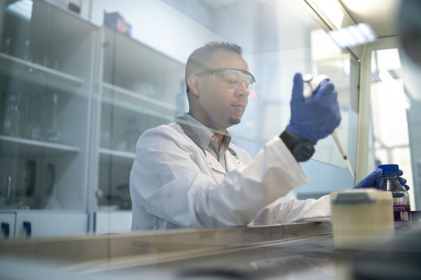 Researcher pipetting inside a fume hood, viewed through the glass sash in cool blue-white light