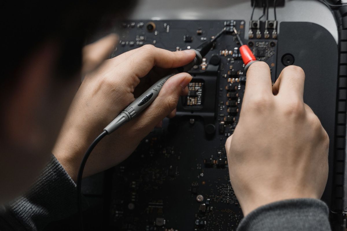 Hands holding two probes against a dense black logic board under low light
