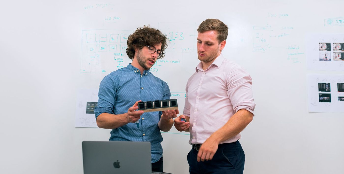 Two engineers examining a sensor array prototype in front of a whiteboard of system diagrams