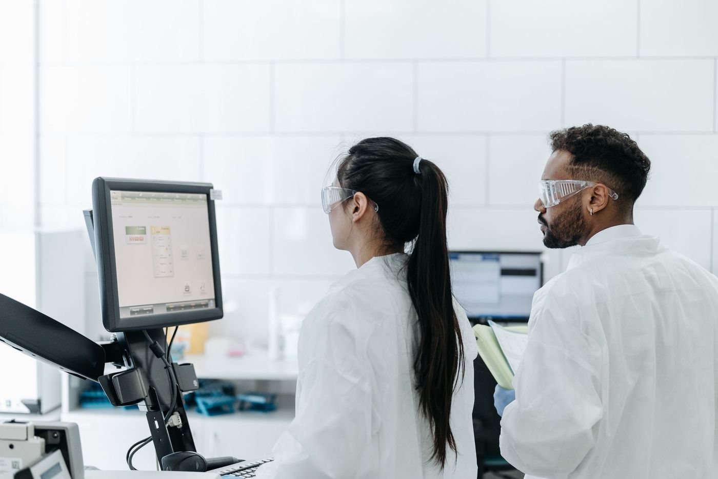 Two researchers in safety glasses reading an instrument control screen together in a white-tiled lab