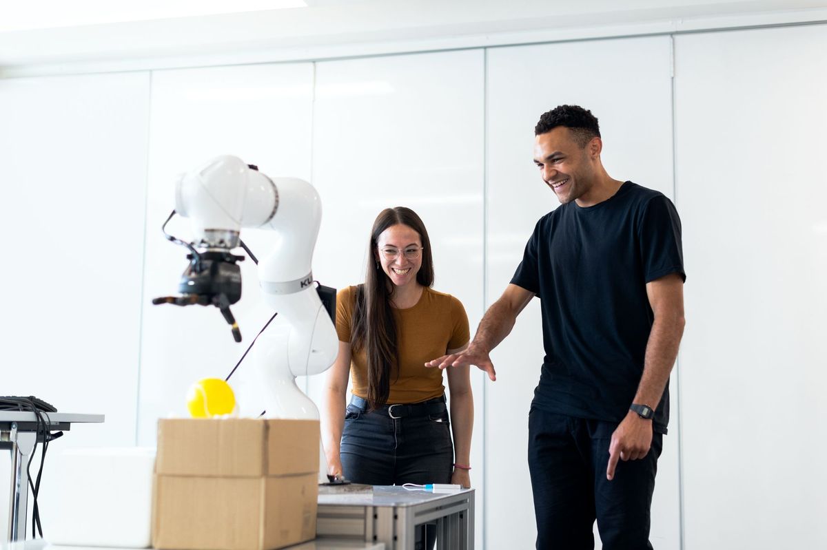 Two engineers in a white research lab watching a collaborative robot arm attempt to pick up a ball
