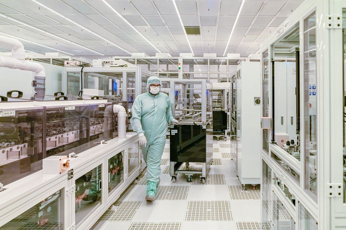Gowned engineer wheeling a wafer cart down a bright aisle of process tools in a semiconductor cleanroom