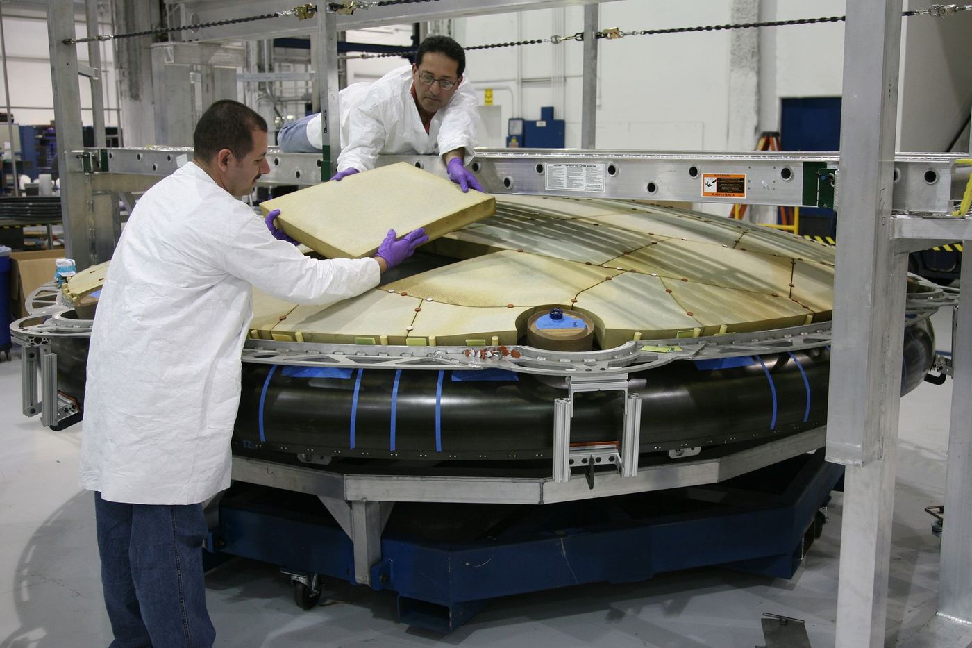 Two technicians in lab coats and purple gloves fitting an insulation panel onto a large spacecraft structure
